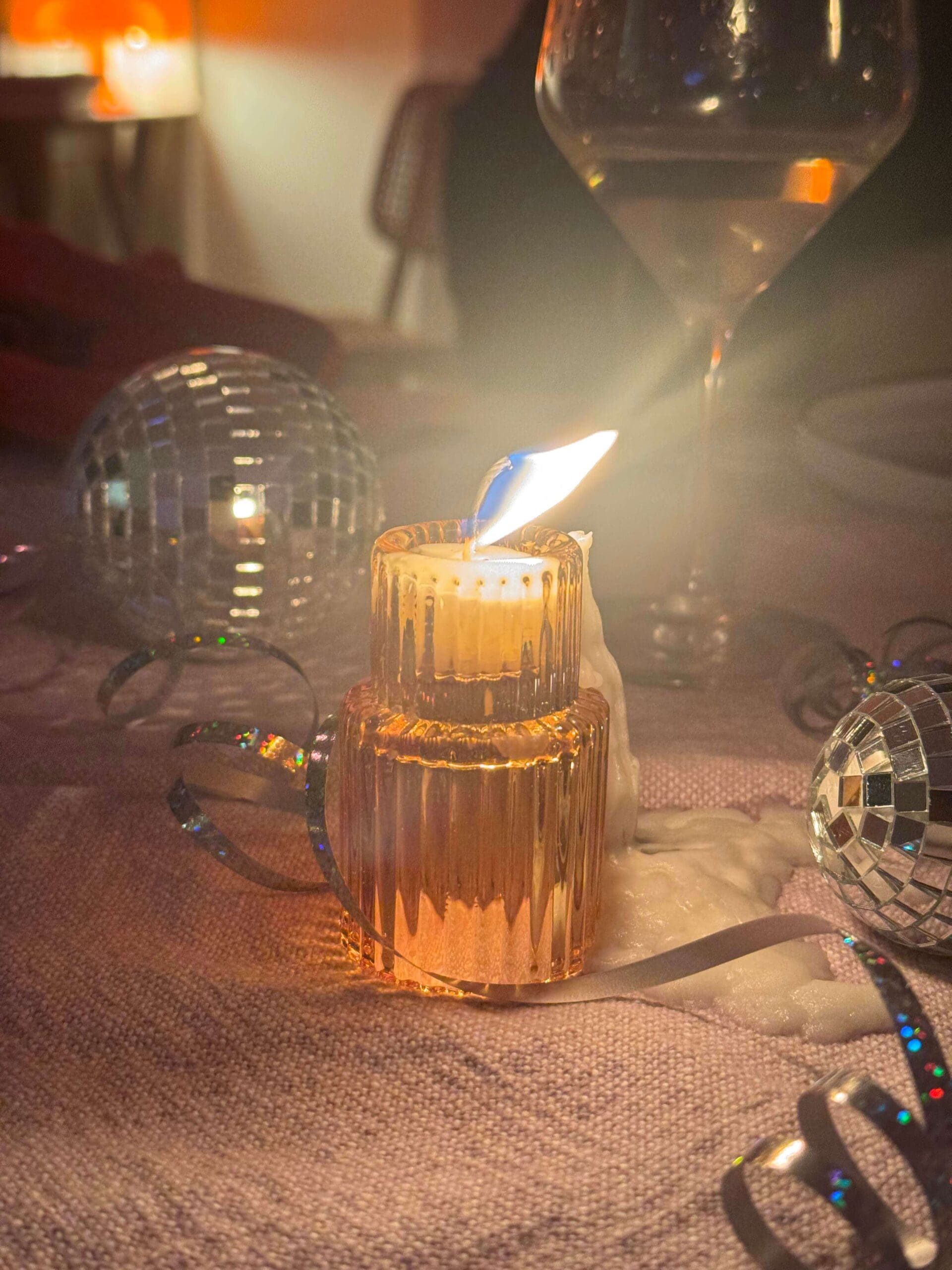 Warm-lit candle in ornate orange glass on a party table with metallic disco balls, shimmering ribbons, spilled wax, and a wine glass