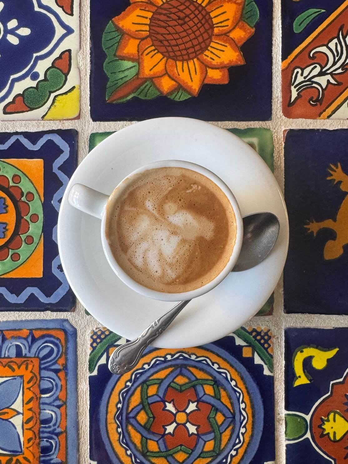 A white cup of coffee topped with latte foam and a spoon on a saucer, placed on colorful patterned talavera tiles featuring floral motifs.
