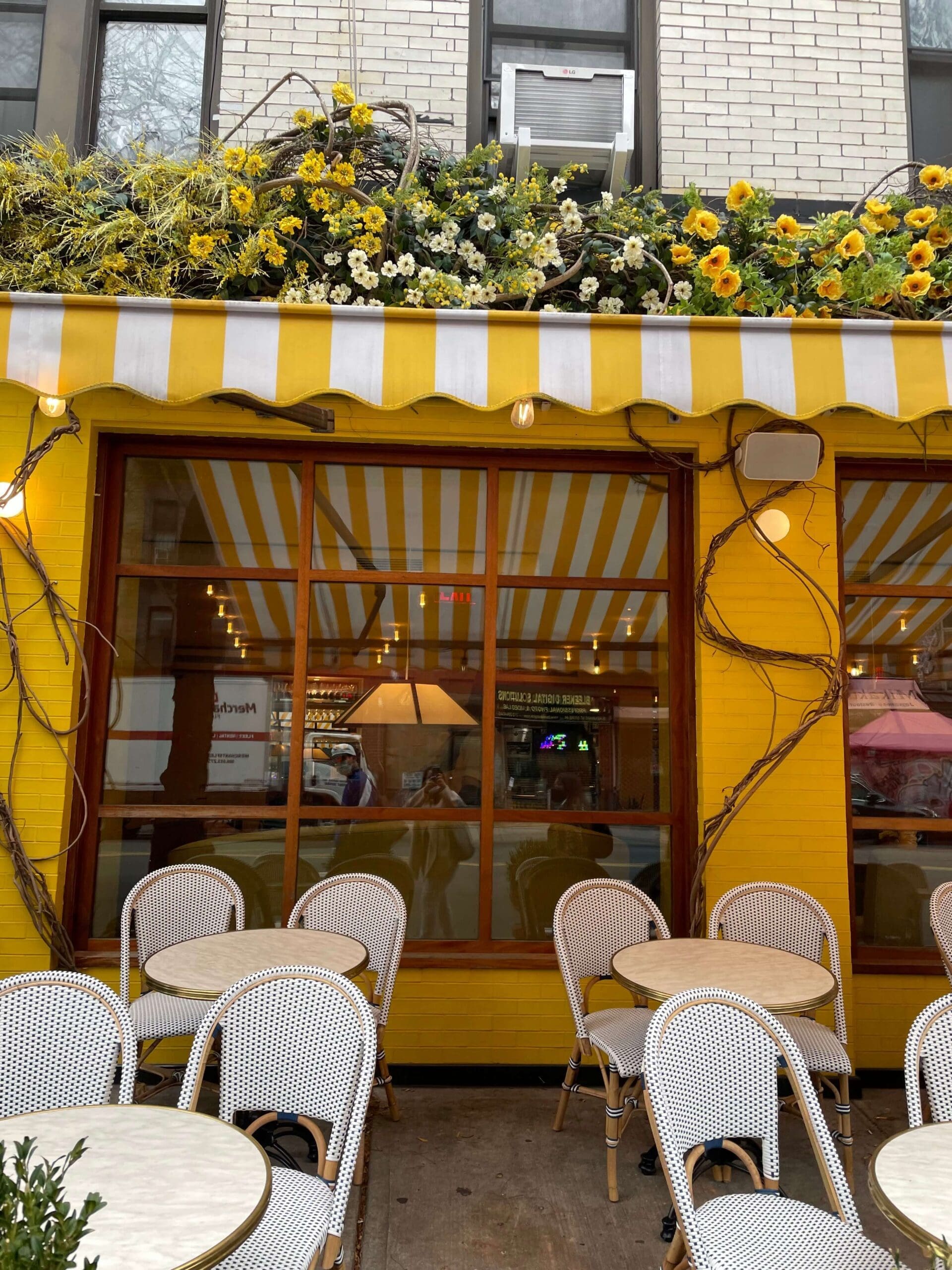 A person takes a selfie in the reflection of a bright yellow cafe window with flowers on the awning.