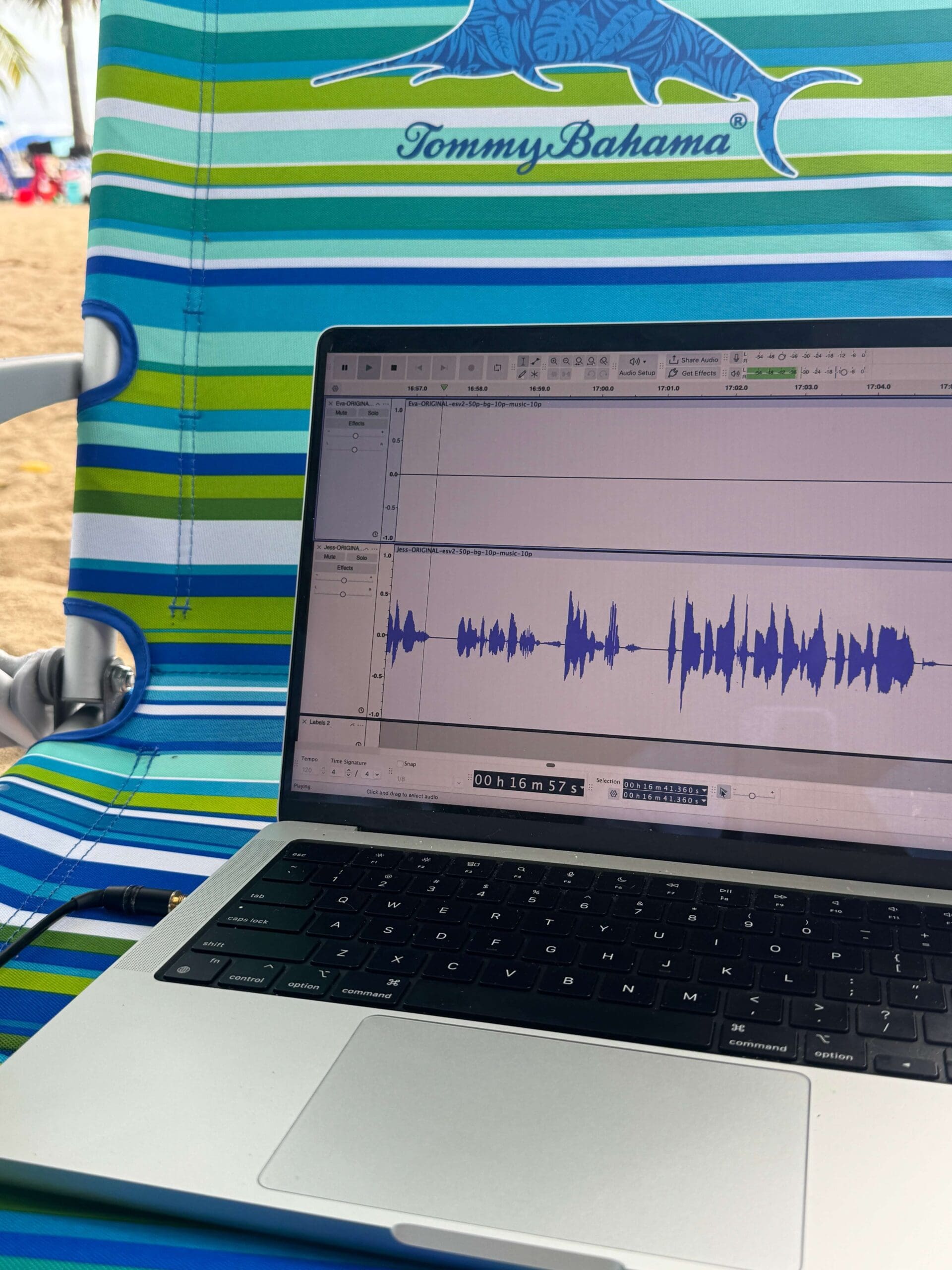 Laptop displaying an audio waveform during podcast editing on a striped beach chair.